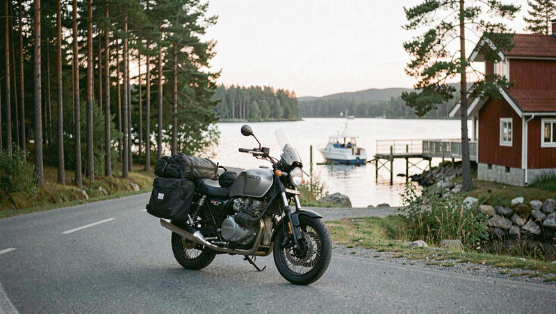 Moto de turismo parada en una carretera rural de Suecia entre bosque de pinos y una casa roja de madera, con lago y embarcadero al fondo.
